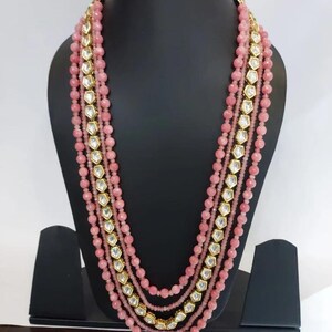 May include: A multi-strand pink beaded necklace with gold accents and clear, geometric-shaped stones. The necklace is displayed on a black mannequin, showcasing its layered design and intricate details. The beads are a soft pink color.