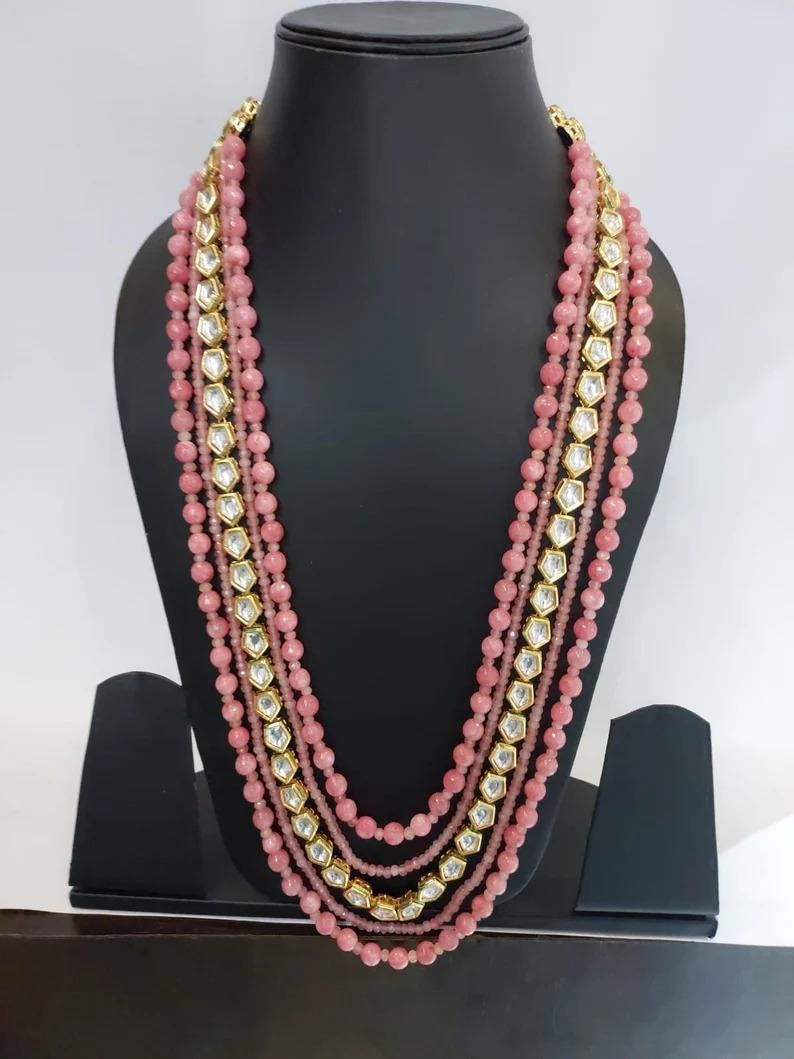 May include: A multi-strand pink beaded necklace with gold accents and clear, geometric-shaped stones. The necklace is displayed on a black mannequin, showcasing its layered design and intricate details. The beads are a soft pink color.