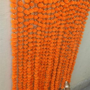 May include: A vibrant orange marigold garland with multiple strands. Each strand features small, round, orange flower heads and silver beads. The garland ends with gold-colored bell-shaped ornaments. Ideal for festive decorations.