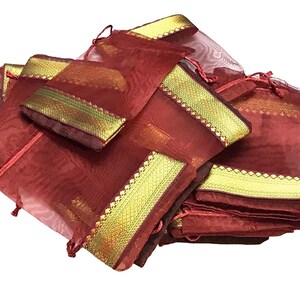 May include: A collection of maroon organza drawstring bags with gold trim. The bags are piled together, showcasing their sheer texture and decorative gold borders. The drawstring closures are a matching maroon color.