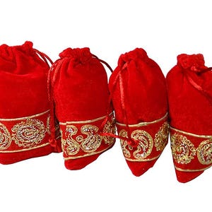 May include: Four red velvet drawstring bags with gold embroidered trim. The bags are closed with matching red drawstrings. The gold trim features an ornate paisley design. The bags are arranged in a row.