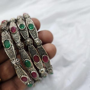 May include: Silver bangles with detailed patterns and alternating red and green gemstones. The bangles are held in a hand, highlighting their ornate design and vibrant colours. These bracelets are a stylish accessory.