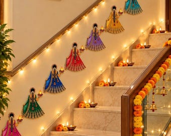 May include: A series of wall-mounted cutouts depicting figures in colourful traditional dresses, each holding a small lit diya lamp. The figures are arranged diagonally along a staircase wall, complemented by string lights and marigold garlands.