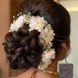 May include: A dark brown updo hairstyle adorned with white flowers and baby's breath. The hair is styled in a bun with loose curls, and the flowers are arranged around the bun. The image is taken from the back, showing the hairstyle and part of the neck.