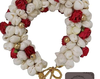 May include: A floral hair accessory featuring white flower buds and red roses, accented with gold beads. The garland is tied with a gold cord. A small, dark gray square with the letters "FWD" in a circular design is in the lower right corner.