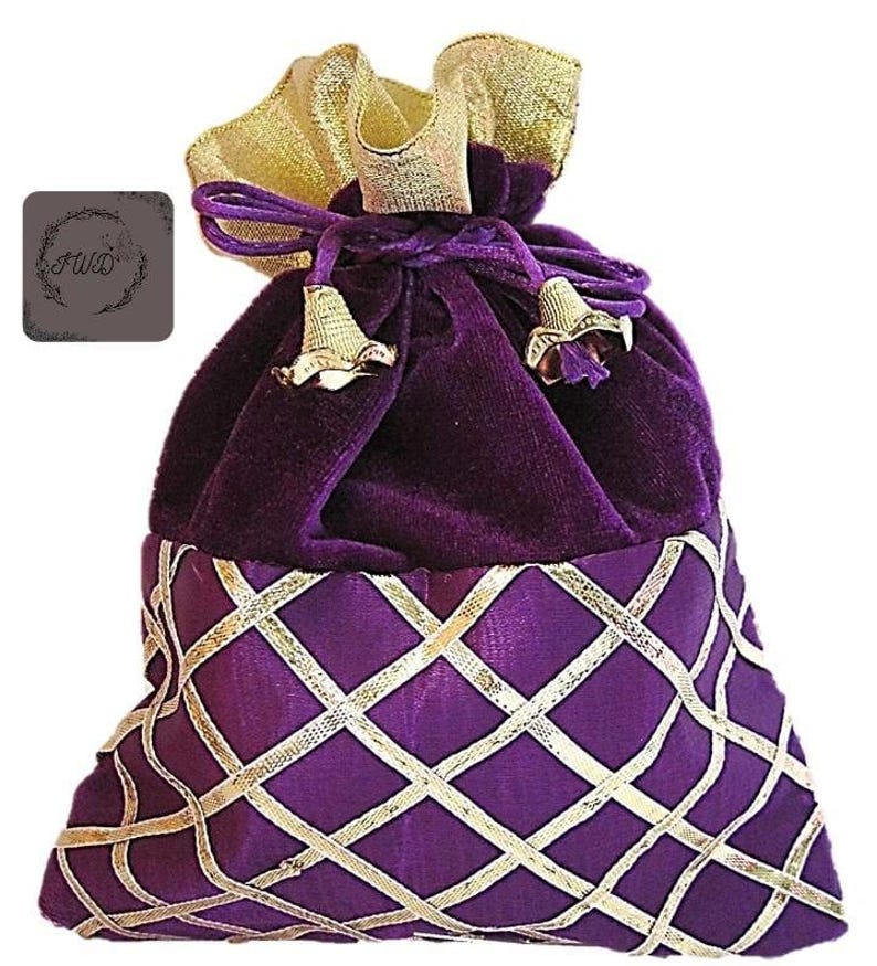 May include: A purple velvet drawstring bag with a gold satin top and gold accents. The lower portion features a purple fabric with a gold diamond pattern. The bag has gold tassels and a small logo on the side.