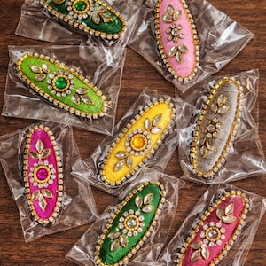 Peut inclure: Plusieurs barrettes ovales colorées dans des emballages transparents. Chaque barrette présente une base en tissu vert, rose, jaune et gris, ornée de perles et d'ornements scintillants. Les barrettes sont présentées sur une surface en bois foncé.