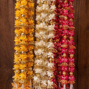 May include: Three decorative floral garlands in yellow, white, and red. Each garland features small flowers, gold-coloured geometric accents, and beaded strands. The garlands hang against a dark wood background.