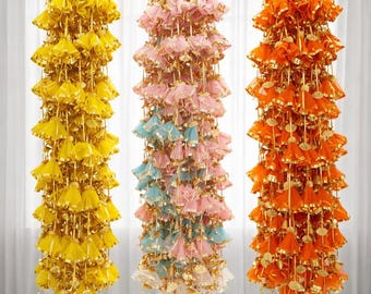 May include: Three hanging decorative garlands in yellow, pink and orange. Each garland is made of layered, bell-shaped fabric with gold trim and hanging gold strands. The garlands are suspended from gold cords.