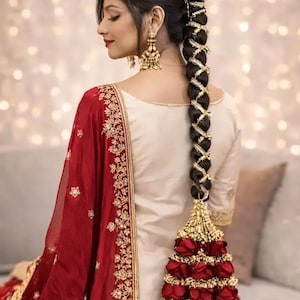 May include: A long dark hair braid adorned with gold ribbon in a crisscross pattern, finished with a large cluster of deep red roses and gold accents. The hair is styled with a gold hair accessory at the top and paired with gold drop earrings.