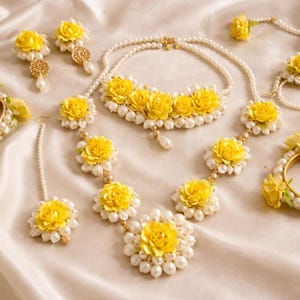 以下が含まれることがあります： 白いサテンの背景に、ネックレス、イヤリング、ブレスレット、ヘアアクセサリーなど、黄色の花のジュエリーのコレクション。ジュエリーは黄色の花と白い真珠が特徴です。