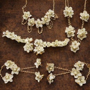 以下が含まれることがあります： アイボリー色の花のジュエリーコレクション。ネックレス、ブレスレット、イヤリング、ヘアアクセサリーが含まれます。繊細な花のモチーフとゴールドトーンのアクセントが特徴で、濃い茶色の木製の表面に配置されています。