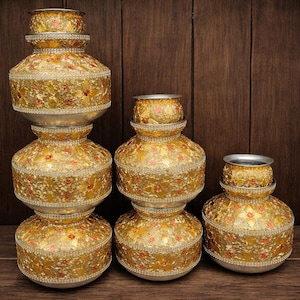 May include: Several decorative gold-coloured metal pots with detailed floral designs and silver rims. The pots, of varying sizes, are arranged on a wooden surface against a dark wood backdrop. These ornate vessels are likely for display purposes.