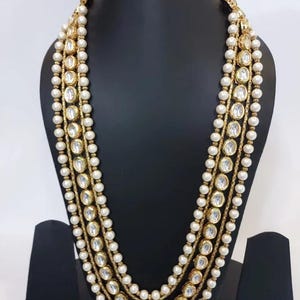 May include: A multi-strand necklace with faux pearls and gold-toned accents. The necklace features large, clear, oval-shaped stones set in gold. The necklace is displayed on a black mannequin.