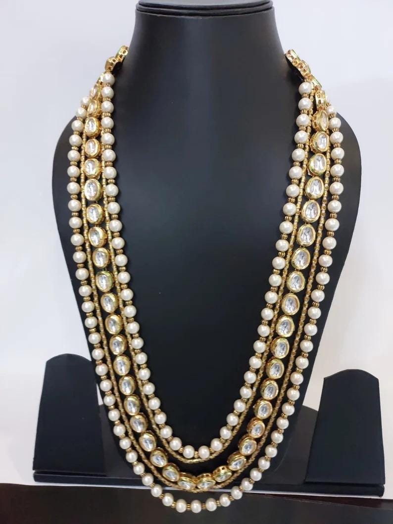 May include: A multi-strand necklace with faux pearls and gold-toned accents. The necklace features large, clear, oval-shaped stones set in gold. The necklace is displayed on a black mannequin.