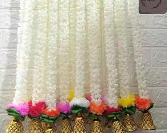 May include: White floral garlands with colorful accents of pink, red, orange, and yellow flowers. Each garland is adorned with gold-colored bell-shaped ornaments at the base. The garlands are hanging against a white brick wall.