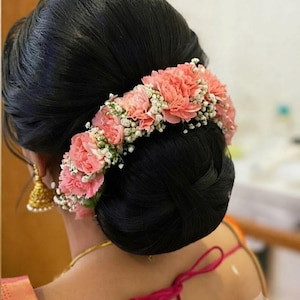 May include: A dark hair bun styled with a floral arrangement featuring soft pink carnations and small white baby's breath flowers. The hair is pulled back into a smooth, rounded bun, with the flowers placed along the top curve of the hairstyle.