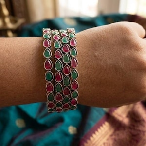 Może przedstawiać: Zestaw trzech srebrzystych bransoletek typu bangle z naprzemiennie ułożonymi czerwonymi i zielonymi kamieniami w kształcie łezki. Każda bransoletka ma powtarzalny wzór tych kolorowych klejnotów osadzonych w metalowej oprawie, tworząc ozdobę.