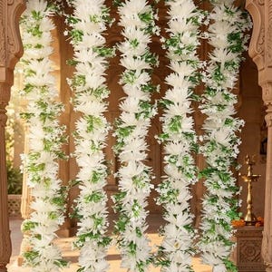 May include: Five vertical garlands of white artificial flowers and green foliage hanging within a carved stone archway. Each garland features dense white petals and small green leaves, terminating in a large, rounded white floral tassel at the base.