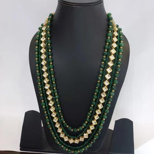 May include: A multi-strand necklace with green beads and gold accents. The necklace features alternating rows of small round beads and larger square, gold-framed, clear stones. The necklace is displayed on a black mannequin.