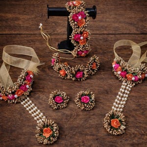 以下が含まれることがあります： ブレスレット、ネックレス、イヤリング、ハンドジュエリーなど、花のジュエリーのコレクション。ピンクとオレンジの花、パール、ゴールドカラーのアクセントが特徴です。ジュエリーは木製の表面に展示されています。