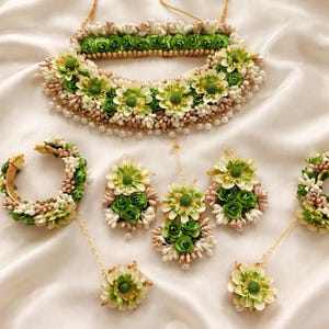 以下が含まれることがあります： ネックレス、ブレスレット、イヤリング、ヘアアクセサリーを含むフローラルジュエリーセット。緑と白の花で飾られ、金と真珠がアクセントになっています。ジュエリーは白いサテンの表面に配置されています。