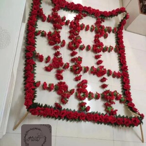 May include: A decorative floral arrangement featuring a white backdrop adorned with vibrant red roses. The roses are arranged in a grid pattern and border the edges, creating a visually appealing display. The arrangement is supported by wooden dowels.