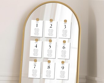 May include: A gold-framed, arched mirror displays nine white table cards, each numbered and held by a gold-colored seal. The cards list names in a simple, elegant font. The mirror reflects a bright, neutral background, suggesting a wedding or event setting.