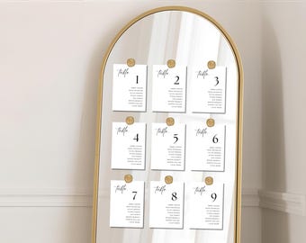 Może przedstawiać: Złota, łukowata rama lustra prezentuje dziewięć białych kart z czarnym tekstem, każda oznaczona jako „table” i ponumerowana od 1 do 9. Karty są przymocowane złotymi pieczęciami woskowymi, prawdopodobnie do planu stołów weselnych.