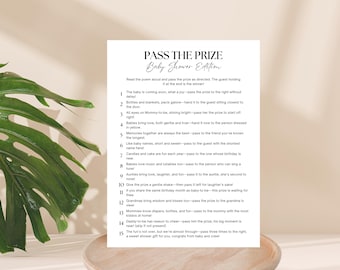 May include: A white baby shower game card titled "PASS THE PRIZE Baby Shower Edition" with a poem. The card is on a light brown surface, with a large green leaf in the background.
