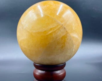 Puede incluir: Una esfera de color amarillo dorado, posiblemente una bola de cristal, se encuentra sobre un soporte de madera marrón oscuro. La esfera tiene una superficie lisa y pulida con sutiles variaciones de color y textura. El soporte tiene un diseño redondeado y escalonado.
