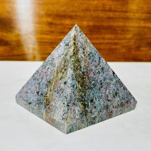 Könnte beinhalten: Eine pyramidenförmige Steinskulptur mit strukturierter Oberfläche. Der Stein weist eine Mischung aus Farben auf, darunter Grau, Grün, Rosa und Blau. Die Pyramide hat eine flache Basis und eine spitze Spitze.