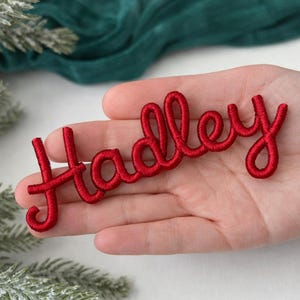 Może przedstawiać: Czerwona, haftowana naszywka z imieniem "Hadley" w kursywie. Naszywka jest trzymana w dłoni, z zieloną tkaniną i gałązkami iglastymi w tle. Imię jest w pogrubionej, pętelkowej czcionce.