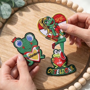May include: Embroidered patches featuring cartoon frogs. One frog holds a heart-shaped patch with the text "Oeteldonk". Another patch shows a frog with a heart that says "I LOVE" and the text "OETELDONK". A third patch shows a frog with the text "SHE 399".