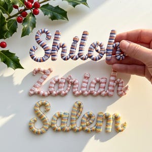 May include: Three yarn-wrapped name signs: "Shiloh" in blue, orange, and white stripes, "Zariah" in pink and white stripes, and "Salem" in yellow, blue, and orange stripes. The signs are displayed on a white surface.