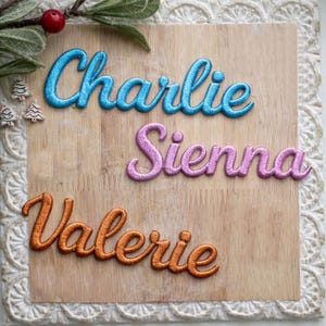 May include: Embroidered names "Charlie", "Sienna", and "Valerie" in blue, pink, and orange thread on a wooden plaque. The plaque is framed with a white, textured border. Decorative sprigs and miniature Christmas trees are in the upper left corner.