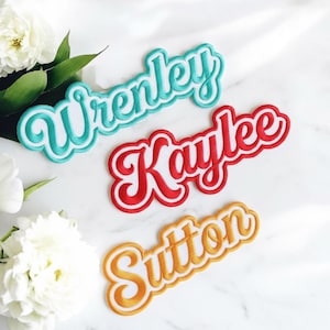 Può includere: Tre toppe ricamate con nomi: "Wrenley" in turchese con contorno bianco, "Kaylee" in rosso con contorno bianco e "Sutton" in arancione con contorno giallo. I nomi sono in corsivo. Le toppe sono su una superficie di marmo bianco.