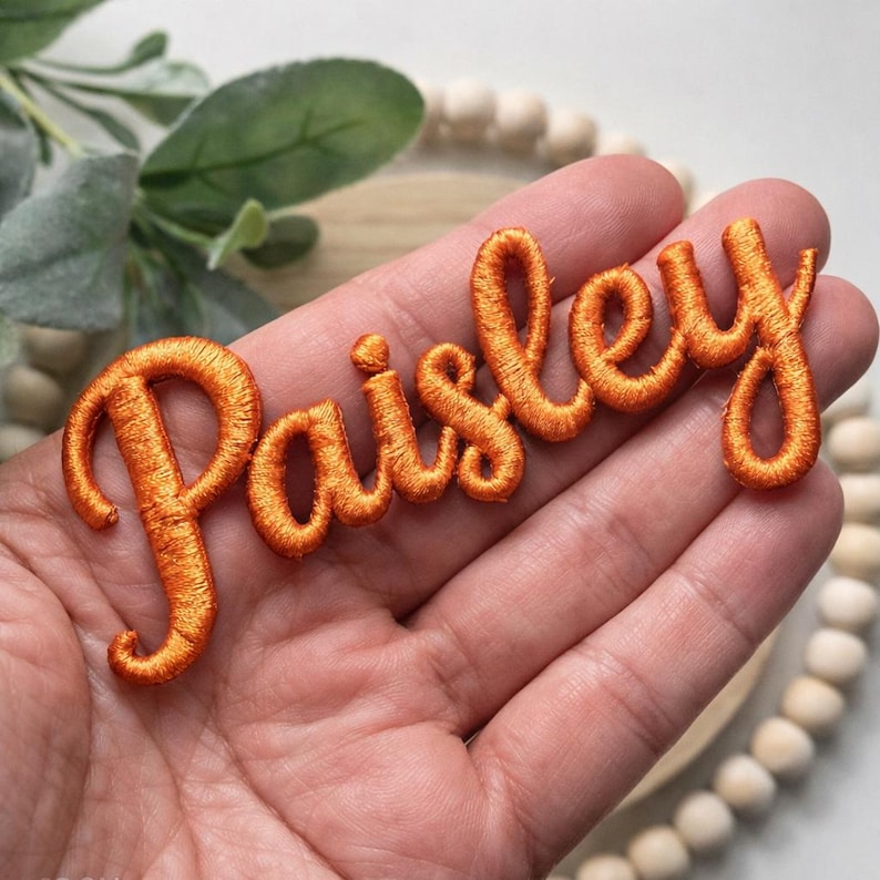 以下が含まれることがあります： オレンジ色の刺繍パッチ。筆記体で「Paisley」と書かれています。パッチは人の手で持たれており、背景にはぼやけた緑と木製のビーズがあります。