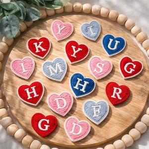 May include: A collection of heart-shaped patches embroidered with letters. The patches are in various colors, including red, pink, and blue, with white stitching and lettering. They are arranged on a wooden tray with a beaded border.