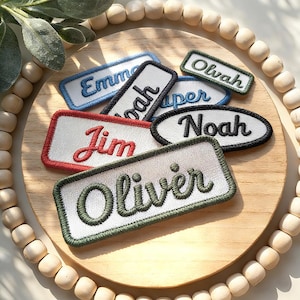 Op de afbeelding: Een verzameling geborduurde naamlabels in verschillende kleuren, waaronder rood, blauw en groen. Elk label heeft een andere naam in een cursief lettertype, met een contrasterende rand. De labels zijn gerangschikt op een houten oppervlak.