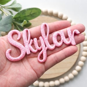 以下が含まれることがあります： 筆記体で「Skylar」と書かれたピンク色の刺繍パッチ。木製の背景と緑が見える手でパッチが持たれています。文字は鮮やかなピンク色です。