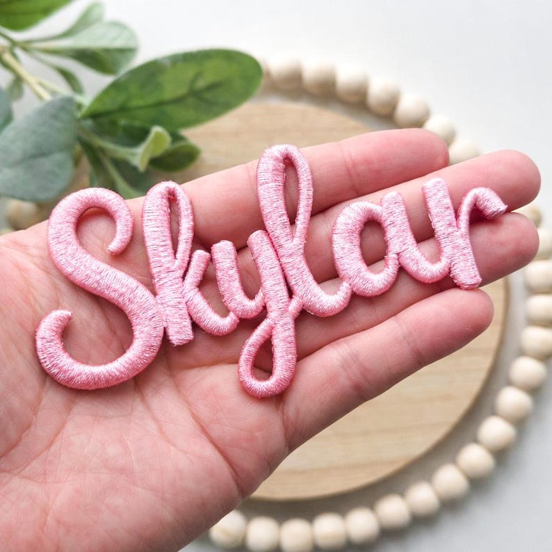 以下が含まれることがあります： 筆記体で「Skylar」と書かれたピンク色の刺繍パッチ。木製の背景と緑が見える手でパッチが持たれています。文字は鮮やかなピンク色です。