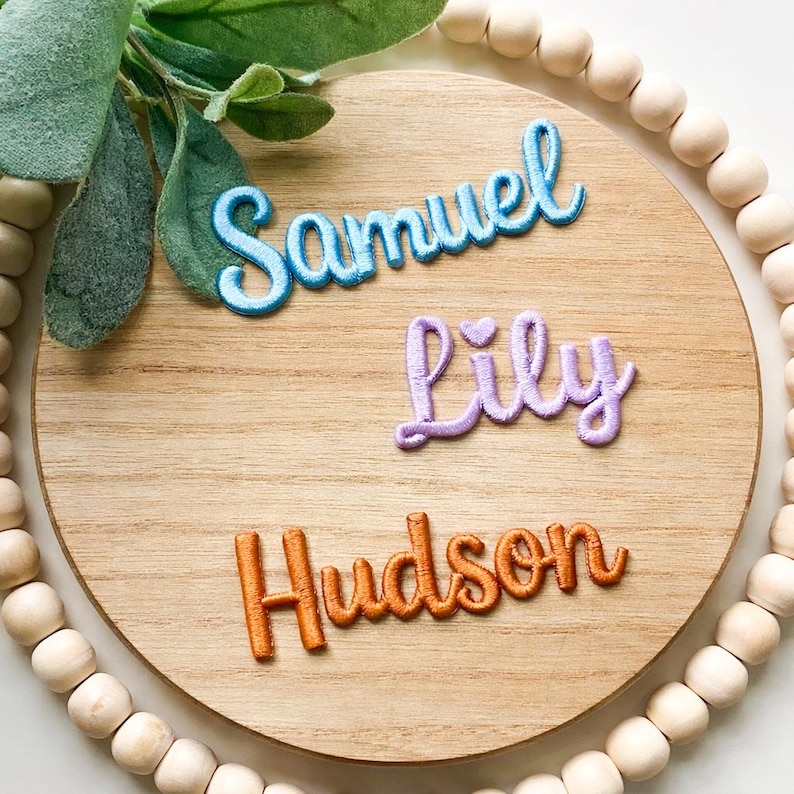 以下が含まれることがあります： 青、紫、オレンジ色の糸で刺繍された「Samuel」、「Lily」、「Hudson」の名前が書かれた木製の丸い看板。看板は緑と木製のビーズで飾られています。