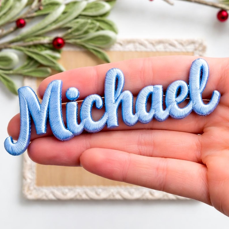以下が含まれることがあります： 「Michael」と書かれた水色の刺繍名札。ピンは人の手で持たれています。ピンは筆記体フォントで、ピンの裏地が付いています。背景には緑と装飾フレームが含まれています。