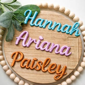 Peut inclure: Panneau rond en bois avec les noms "Hannah", "Ariana" et "Paisley" brodés en fil bleu, violet et orange. Le panneau est orné de perles en bois et de verdure.