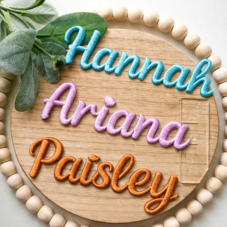 以下が含まれることがあります： 青、紫、オレンジ色の糸で刺繍された「Hannah」、「Ariana」、「Paisley」という名前が書かれた木の丸い看板。 看板は木製のビーズと緑で飾られています。