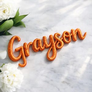 Può includere: Nome "Grayson" ricamato in arancione su una superficie di marmo, con fiori bianchi e foglie verdi sullo sfondo. Il nome è in corsivo, ideale per decorazioni personalizzate.