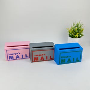 May include: Three rectangular mail organizers in pink, gray, and blue. Each box features a horizontal slot at the top and a rectangular border containing the text "Lauryn's MAIL," "Joseph's MAIL," and "Elijah's MAIL" in matching colored lettering.