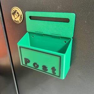 May include: A bright green plastic wall-mounted mailbox featuring a rectangular slot and a hinged lid. The front panel displays the word Post in raised black lettering. The container is attached to a dark surface, with a small circular gold emblem.