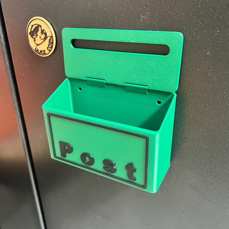 May include: A bright green plastic wall-mounted mailbox featuring a rectangular slot and a hinged lid. The front panel displays the word Post in raised black lettering. The container is attached to a dark surface, with a small circular gold emblem.